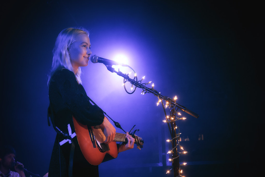 Phoebe Bridgers singing into a microphone whilst playing the guitar