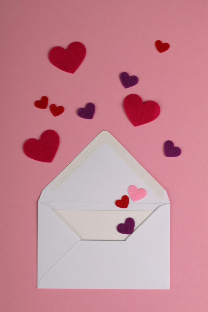 An open white envelope laid flat against a pink background. Emerging from the envelope are some carefully-cut pink and purple paper hearts. The image is used to reflect the love and emotion that Sabrina Carpenter put into her album, based on emails she sent to herself.