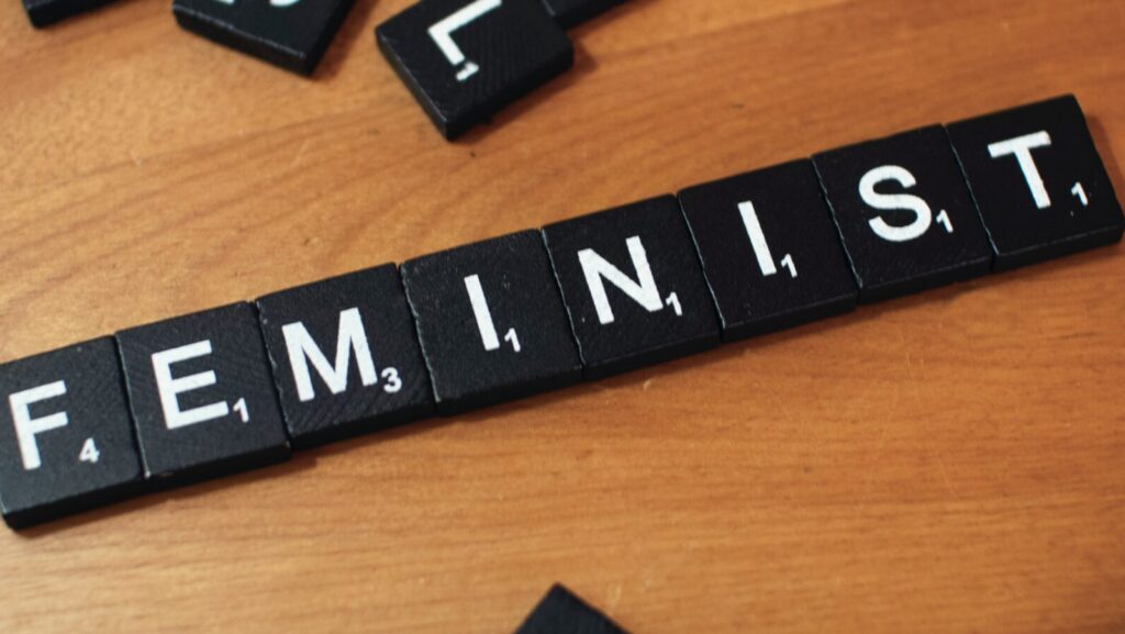 The word 'feminist' spelled out using black and white Scrabble tiles on a wooden surface.