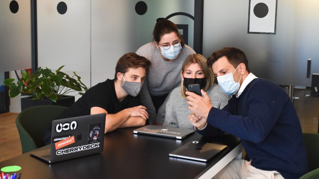 Four people looking at a phone screen. They all wear a mask.