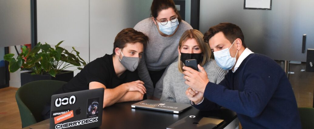 Four people looking at a phone screen. They all wear a mask.