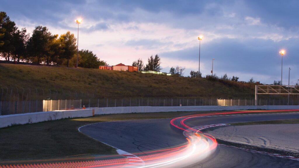 racing track at twilight, as the lights start to come on. a red blur is seen to show a car speeding past. formula one strategy is key to ensure high performance in all conditions.