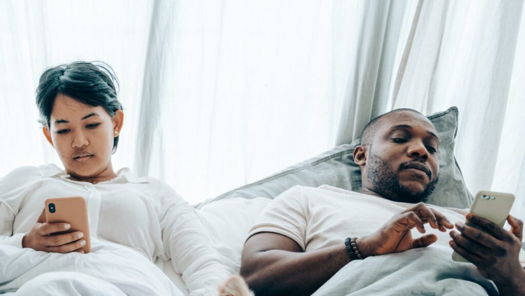 Image shows a woman and a man lying next to each other in bed on their mobile phones