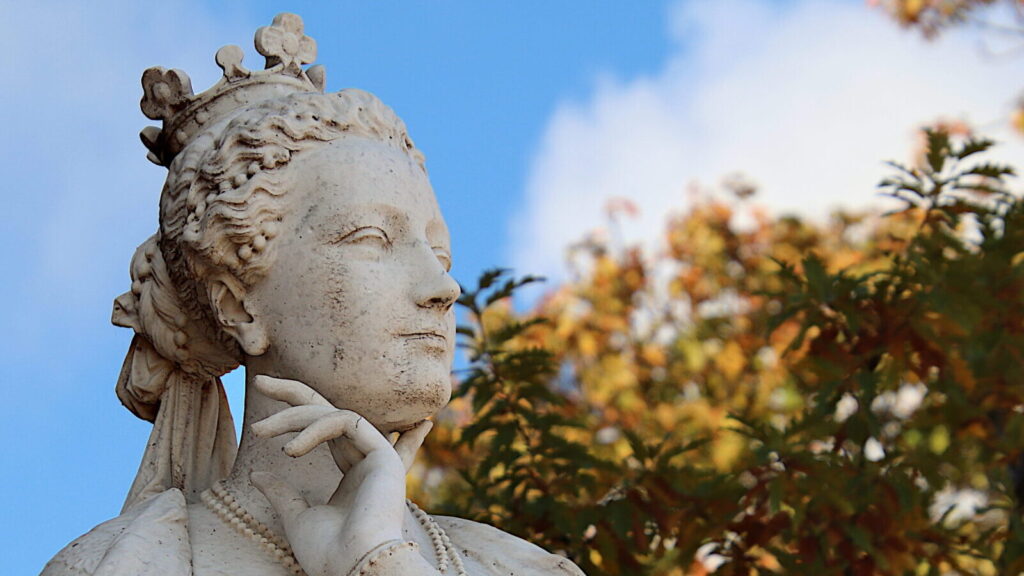 A stone, female statue stands under a blue sky, a hand placed thoughtfully under her chin.