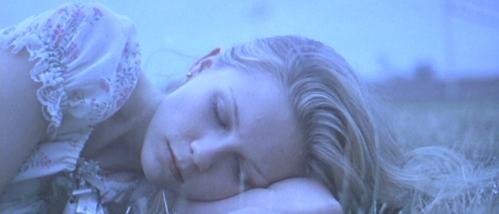 A close up of a blond girl's face as she lies in some grass with her eyes shut.