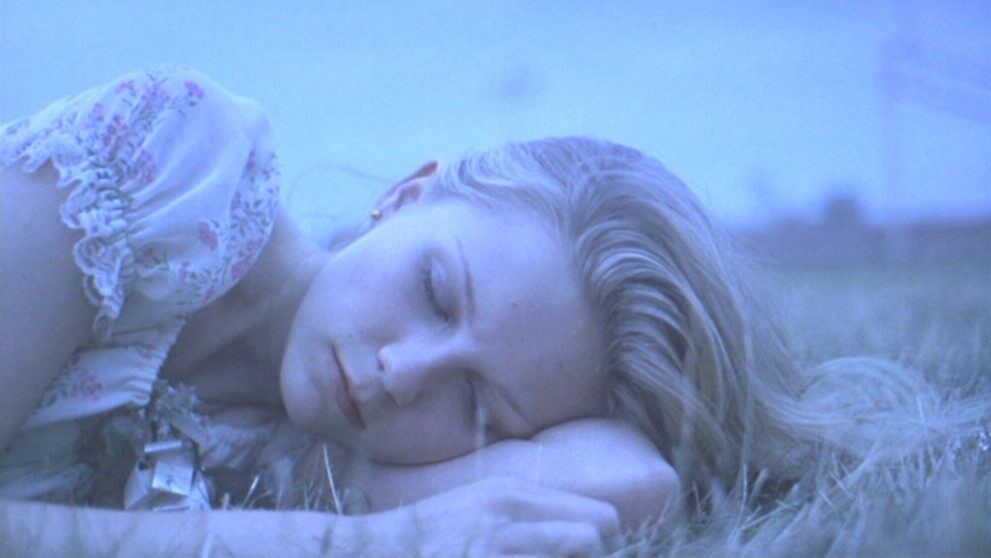 A close up of a blond girl's face as she lies in some grass with her eyes shut.