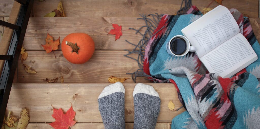 Feet in grey socks standing on stairs surrounded by autumnal leaves, a book, and a pumpkin.