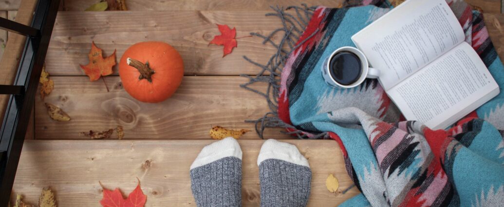 Feet in grey socks standing on stairs surrounded by autumnal leaves, a book, and a pumpkin.