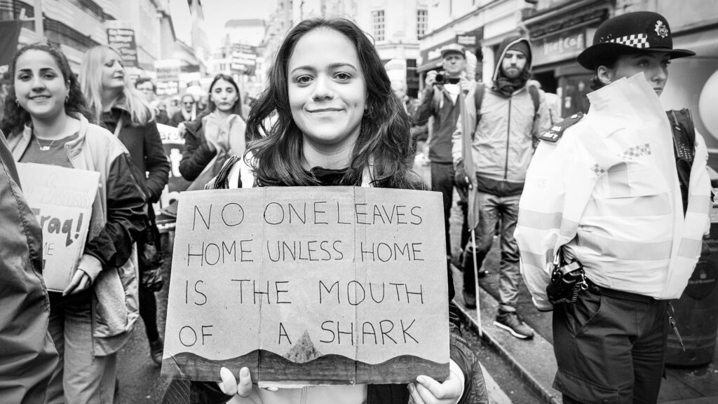 Image taken during the March Against Racism in central London on Saturday on 18 March. A protestor holds a sign reading 'No One Leaves Home Unless Home Is The Mouth Of A Shark'.