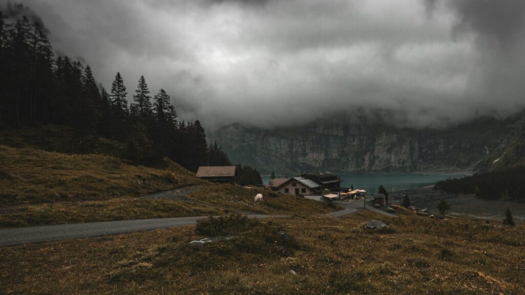 An eerie small town set into a mountainous area surrounded by trees and dark clouds, with a lake in the background.