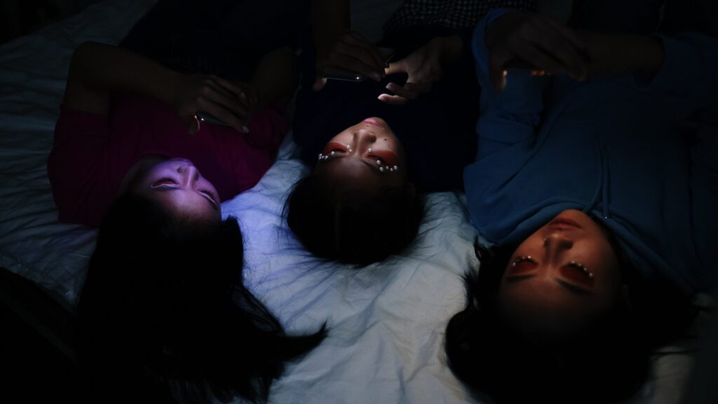 Image shows three women in ethereal 'Euphoria-esque' makeup lying on a bed, their faces lit by their phone screens.