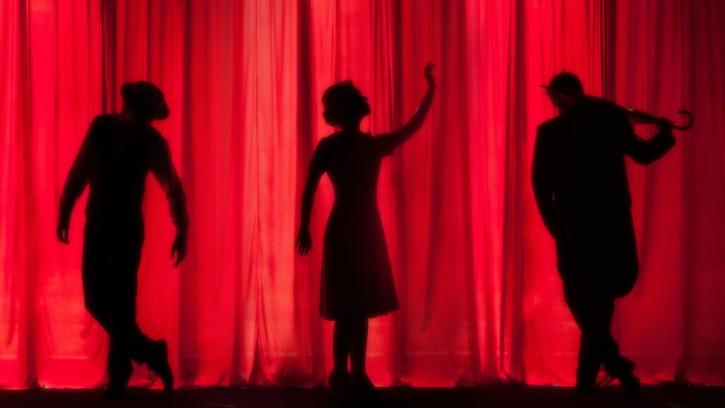 Three silhouettes standing in front of a red stage curtain.