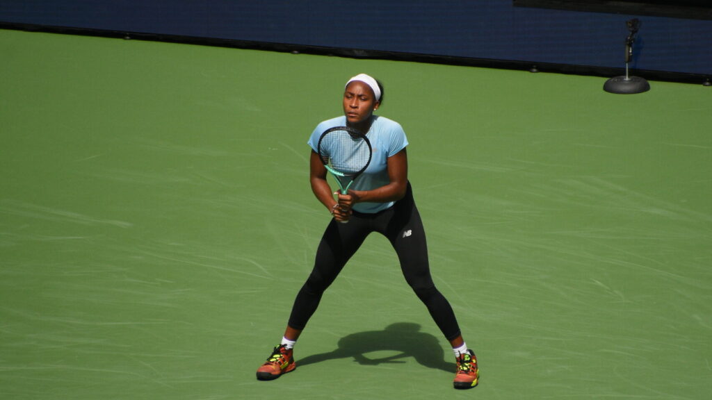 Coco Gauff on a tennis court wearing a blue t-shirt and black sports leggings.