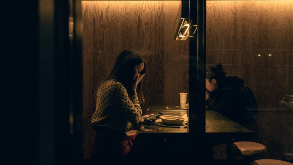 A girl sat at a restaurant table looks at her phone in shock.