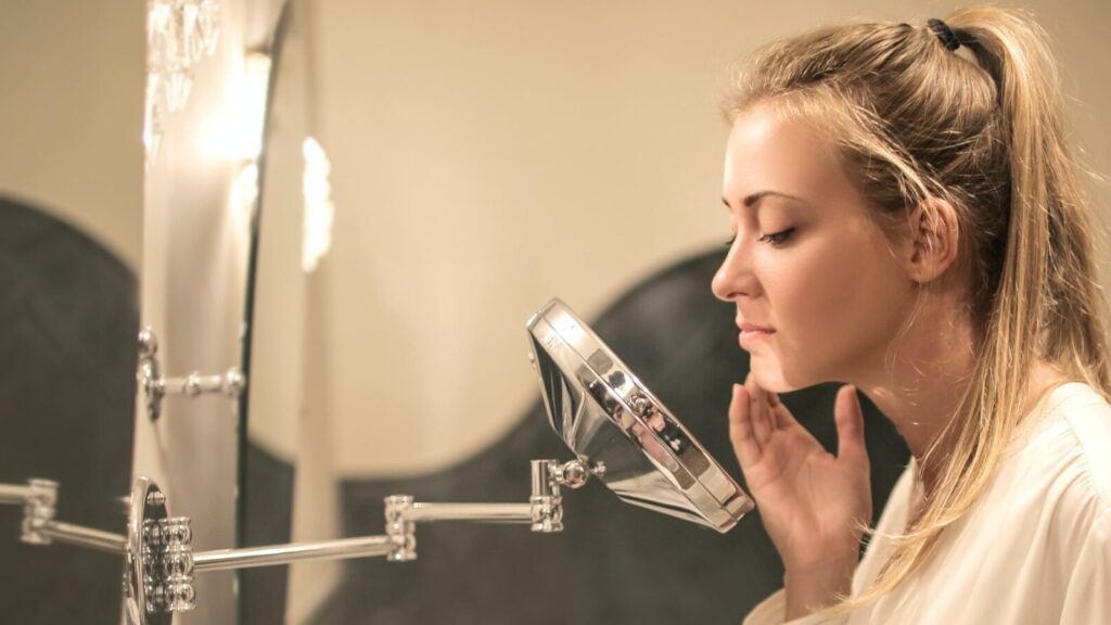 Image shows a woman examining her acne in a bathroom mirror