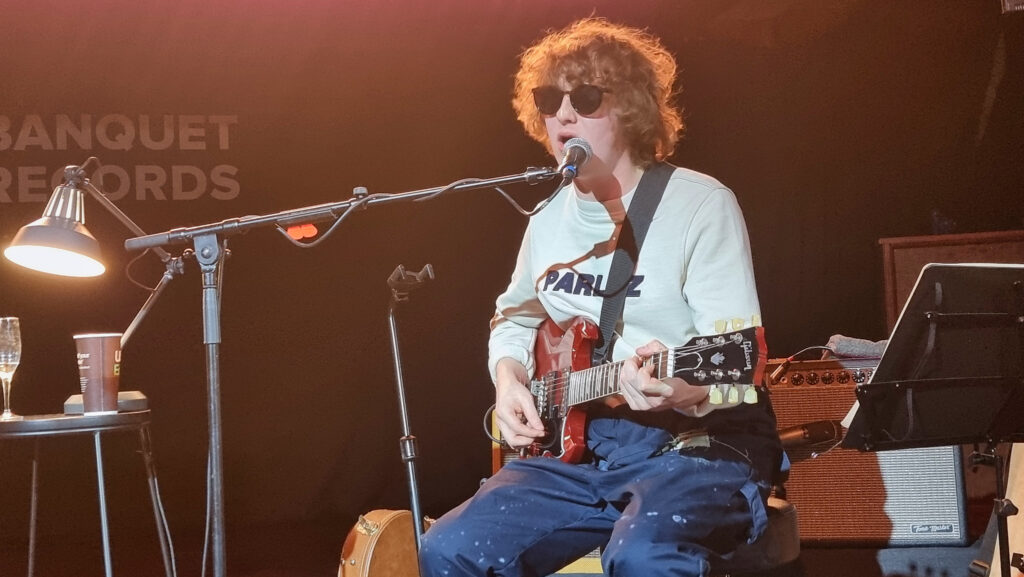 The Snuts member sat on stage wearing a white t-shirt, navy trousers and black sun glasses playing guitar and singing into a microphone.