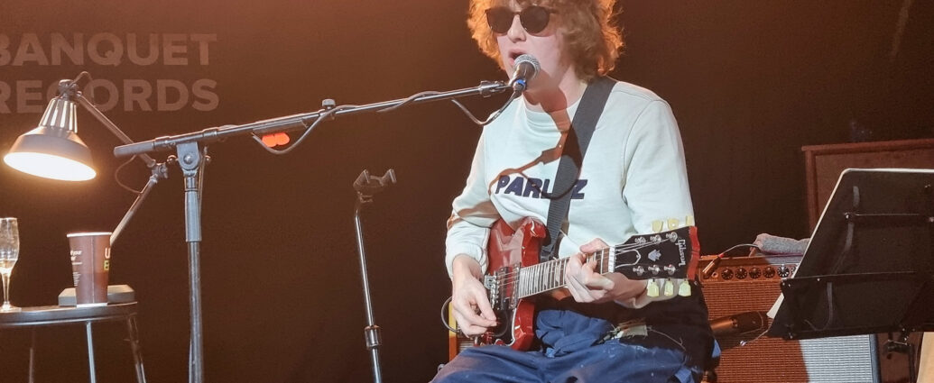 The Snuts member sat on stage wearing a white t-shirt, navy trousers and black sun glasses playing guitar and singing into a microphone.