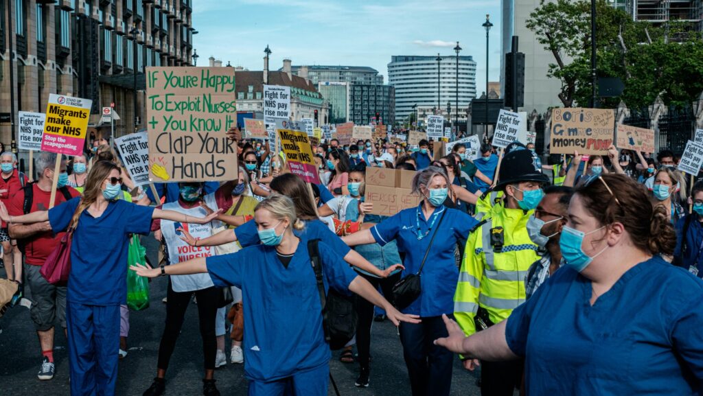 Nurses in blue scrubs and face masks standing with their arms outstretched in the middle of the road during NHS strikes