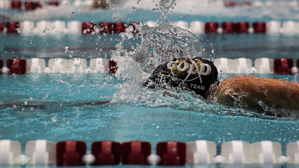 A person wearing a black swimming cap making a splash as they swim along a swimming pool lane alongside red and white floating markers. Achieng Ajulu-Bushell