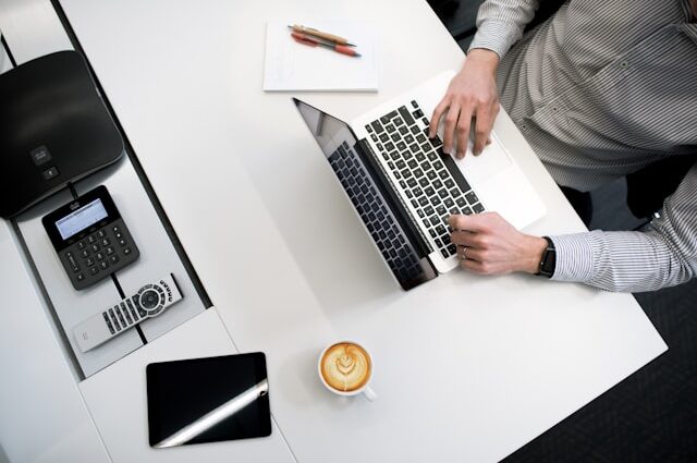 A man in a shirt working at a laptop at desk with coffee, iPad and a notebook alongside. A Journalist's Code of Ethics