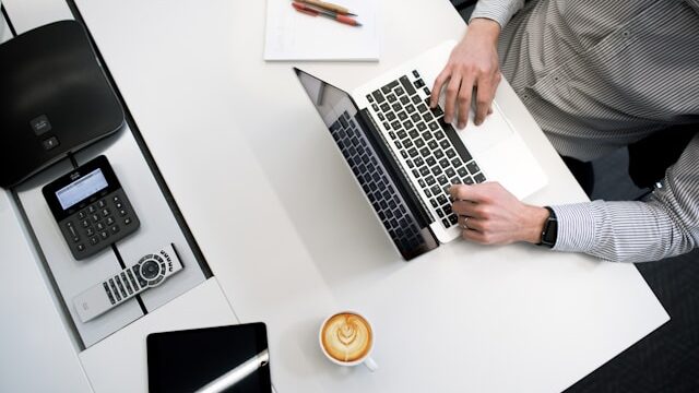 A man in a shirt working at a laptop at desk with coffee, iPad and a notebook alongside. A Journalist's Code of Ethics