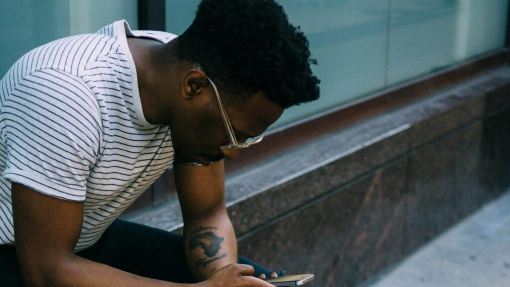 a black man sitting leaning over his mobile phone, reading reclaimed slurs.