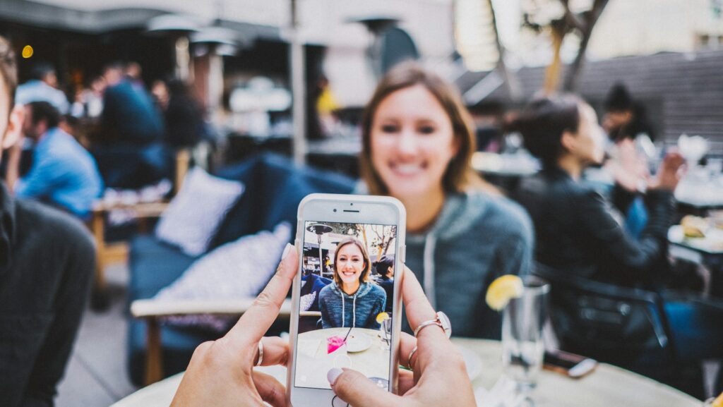 A woman sat at an outdoor restaurant table being photographed on an iPhone.