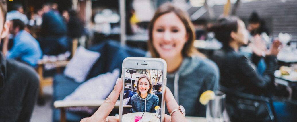 A woman sat at an outdoor restaurant table being photographed on an iPhone.