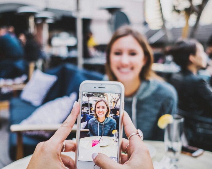 A woman sat at an outdoor restaurant table being photographed on an iPhone.