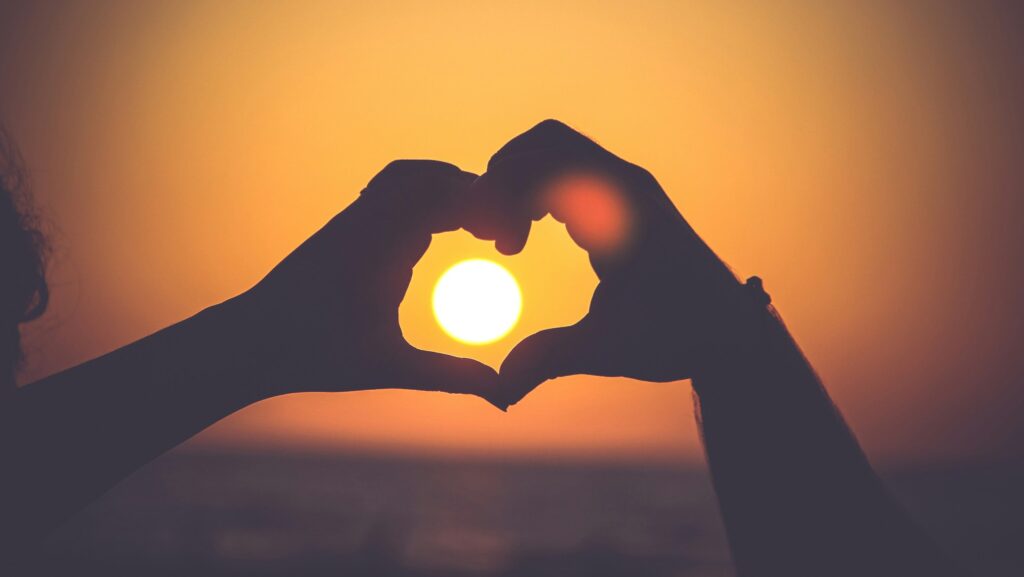 person forming a heart with their hands around a setting sun, backed by amber sky.