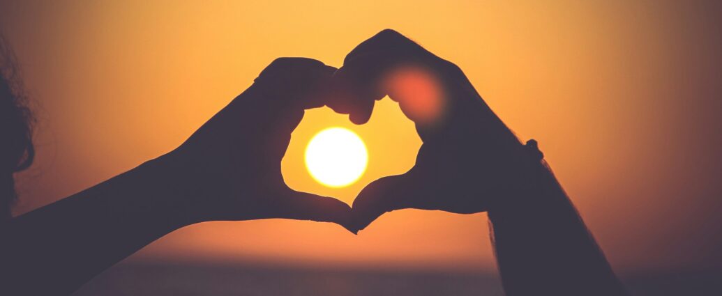 person forming a heart with their hands around a setting sun, backed by amber sky.