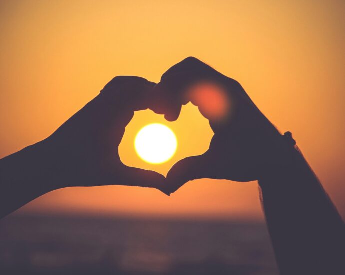 person forming a heart with their hands around a setting sun, backed by amber sky.