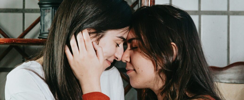 two women, one in white long sleeve top, other in orange long sleeve top, with noses touching and eyes closed.