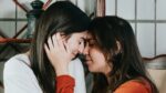 two women, one in white long sleeve top, other in orange long sleeve top, with noses touching and eyes closed.