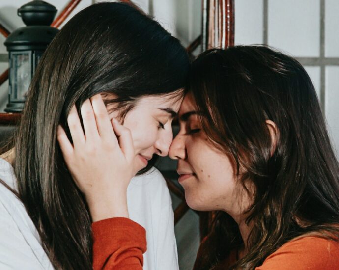 two women, one in white long sleeve top, other in orange long sleeve top, with noses touching and eyes closed.