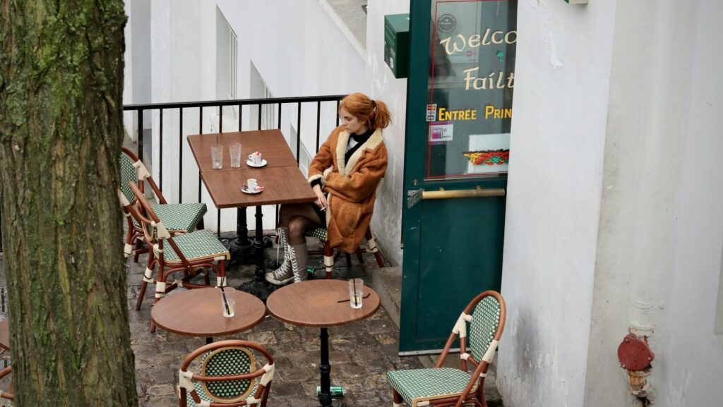 A woman with ginger hair and brown coat sitting at a table outside a restaurant.