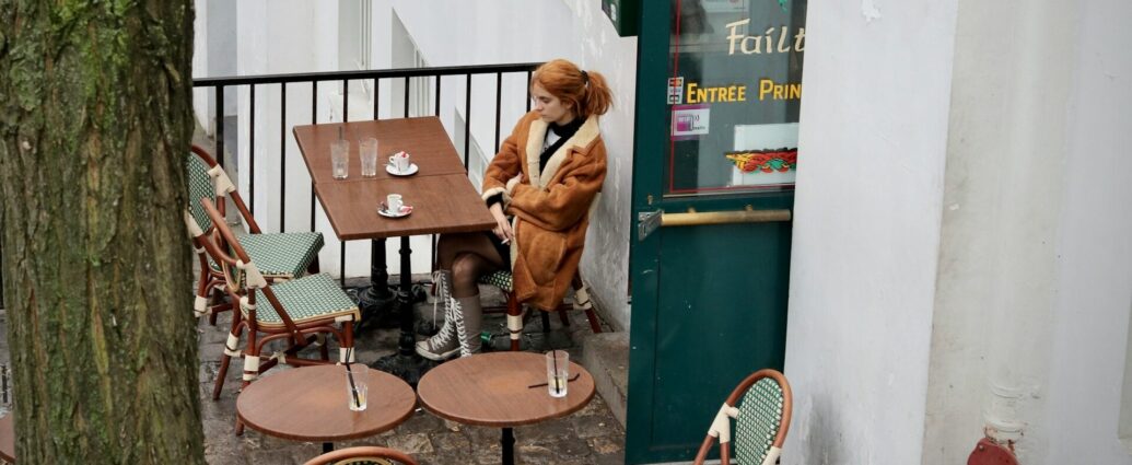 A woman with ginger hair and brown coat sitting at a table outside a restaurant.