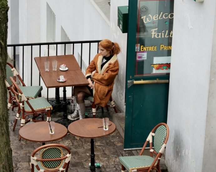 A woman with ginger hair and brown coat sitting at a table outside a restaurant.