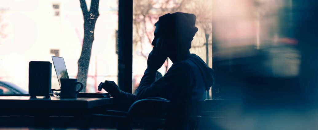 Silhouette of a man wearing a beanie sat in cafe by window on laptop.