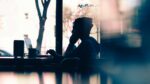 Silhouette of a man wearing a beanie sat in cafe by window on laptop.