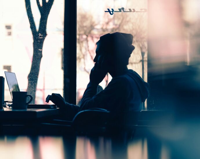 Silhouette of a man wearing a beanie sat in cafe by window on laptop.