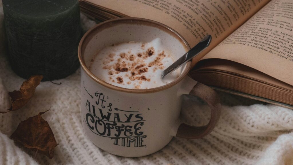 a mug of frothy coffee next to book on cream blanket.