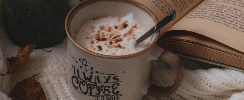 a mug of frothy coffee next to book on cream blanket.