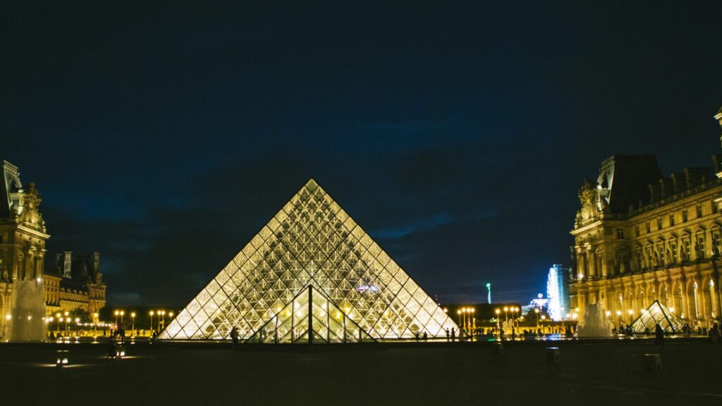 Image of triangular Louvre in Paris lit up in dark. Thousands of people protest in support of Gisèle Pelicot.