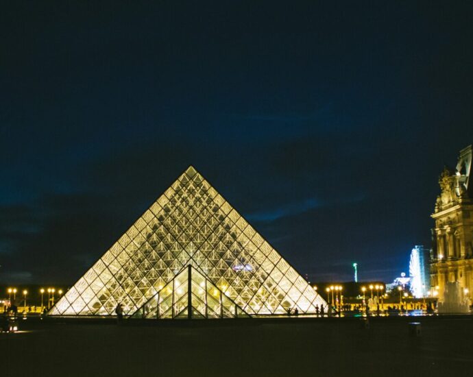 Image of triangular Louvre in Paris lit up in dark. Thousands of people protest in support of Gisèle Pelicot.