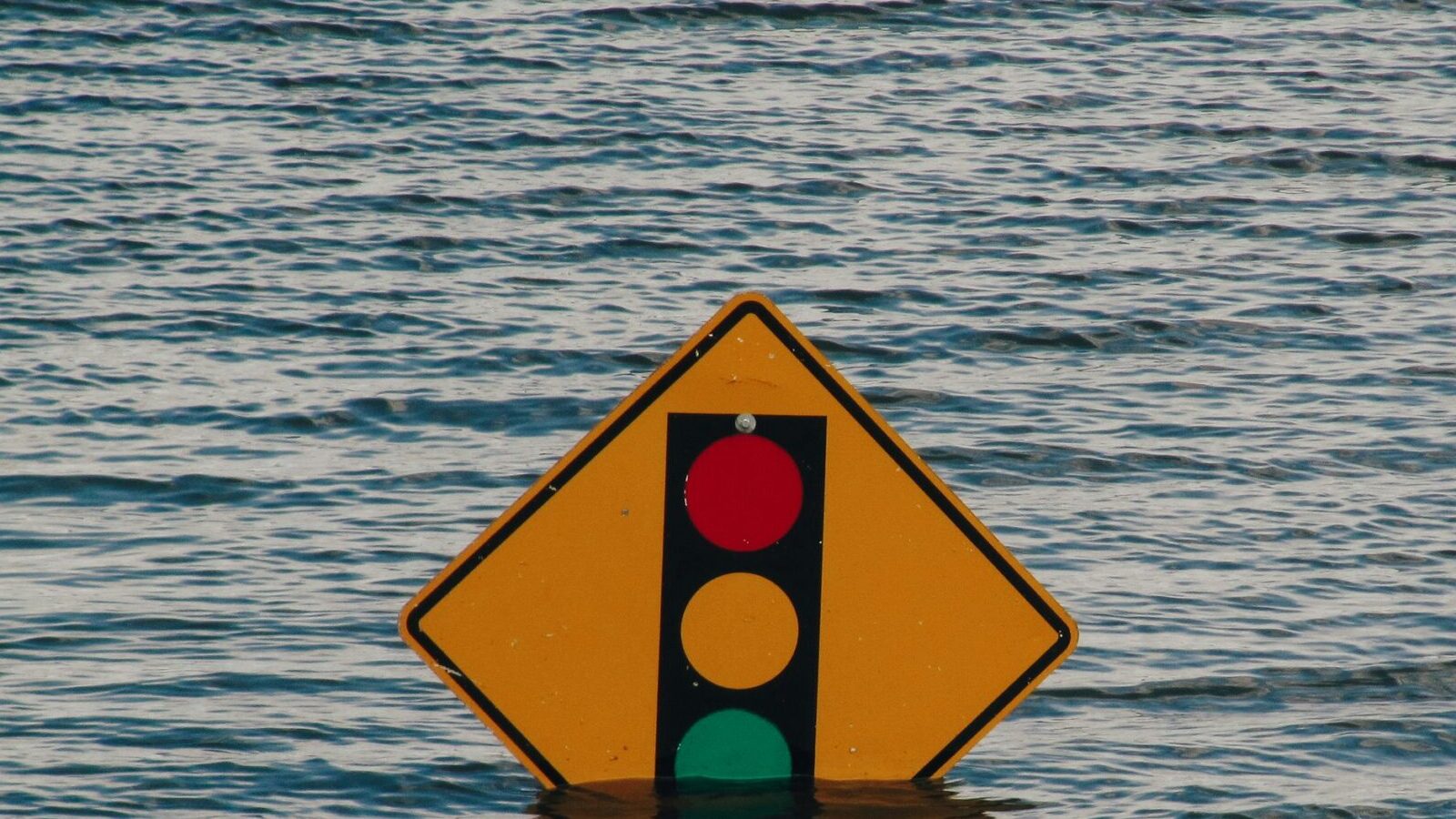 Red, amber and green traffic light symbol on yellow diamond sign sinking in water.