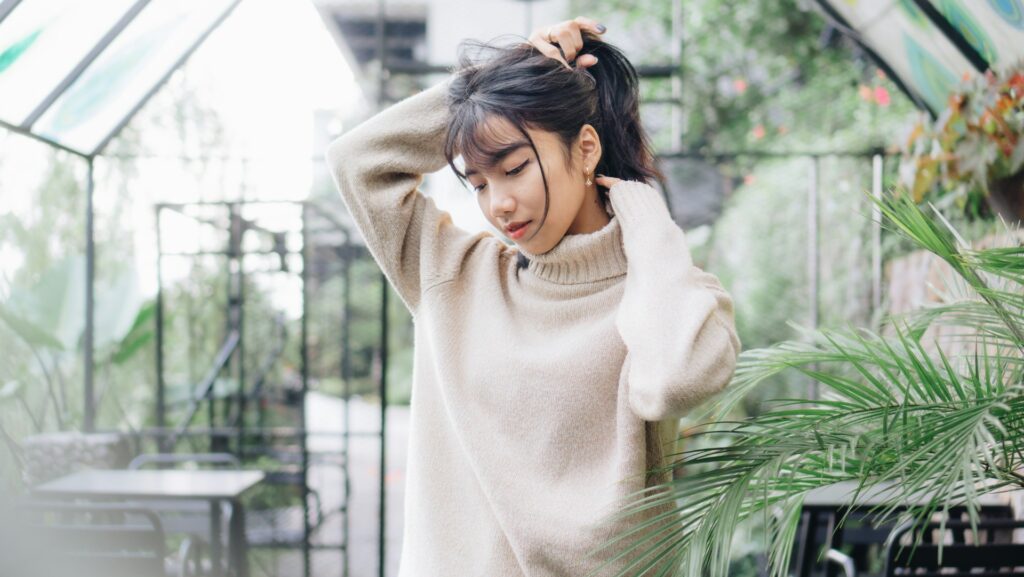 A woman wearing a beige jumper standing in greenhouse holding up her dark brown hair