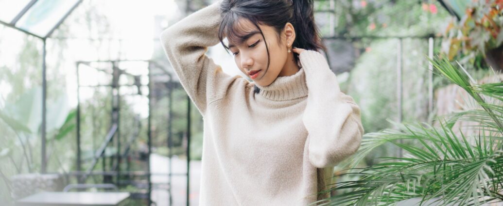 A woman wearing a beige jumper standing in greenhouse holding up her dark brown hair