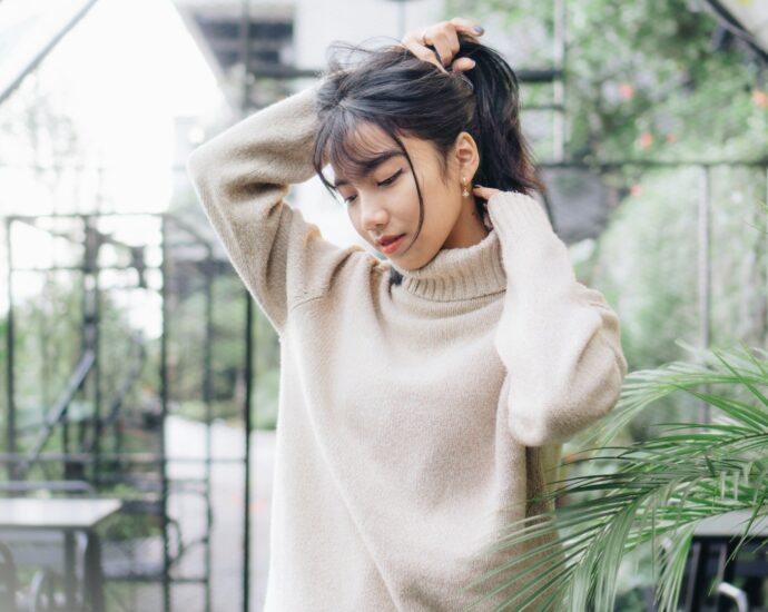 A woman wearing a beige jumper standing in greenhouse holding up her dark brown hair