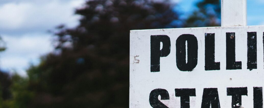 A sign reading 'polling station' in bold black letters, backed by trees and cloudy sky representing 2024 election.